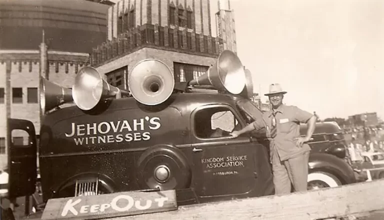 A vintage vehicle marked 'Jehovah's Witnesses' with multiple loudspeakers mounted on top, parked beside a wooden barrier.