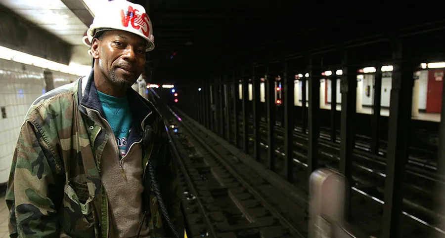 A subway worker wearing a hard hat and camouflage jacket stands near train tracks in a subway station.