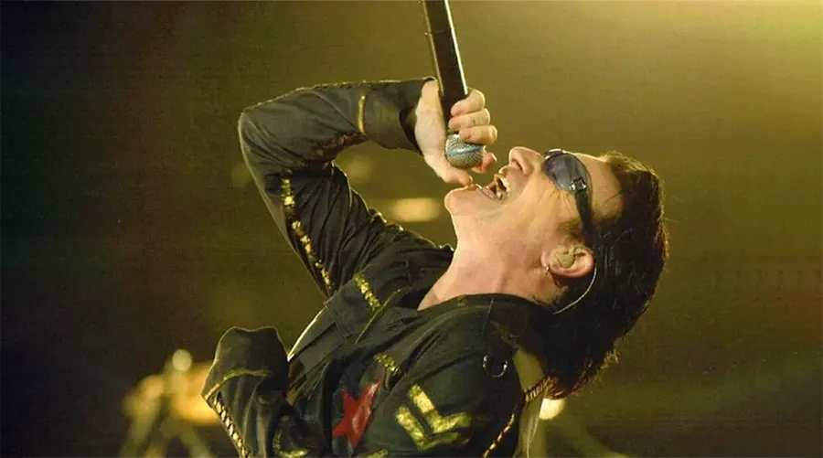 Bono singing into a microphone while wearing sunglasses and a black jacket during a live performance.