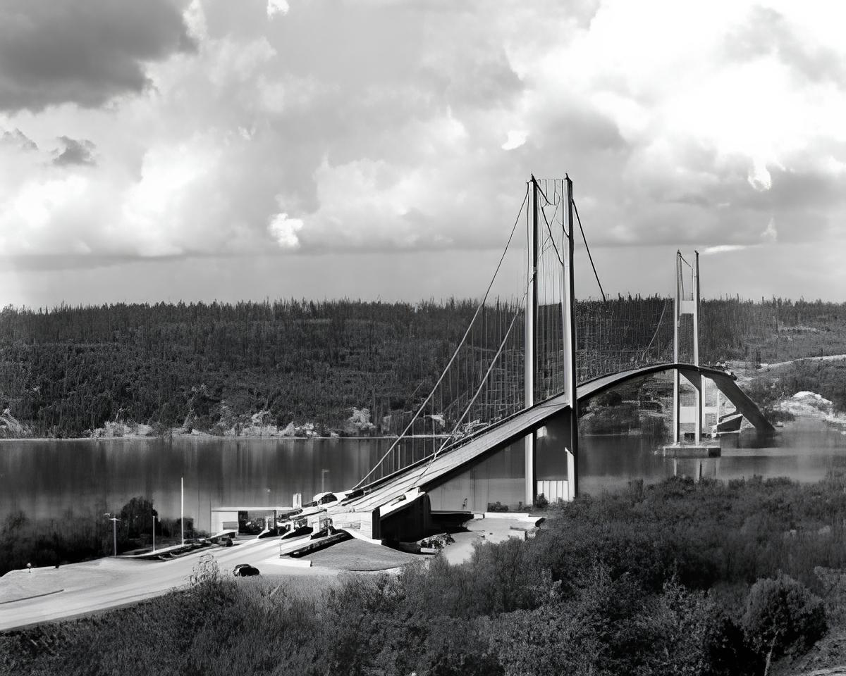 Black and white image of a suspension bridge spanning over a body of water with forested hills in the background.