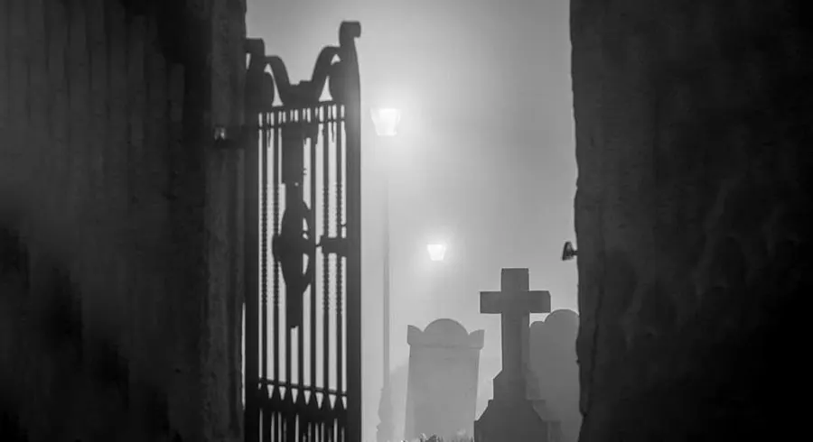 Foggy cemetery scene featuring a cross and a wrought iron gate in black and white.