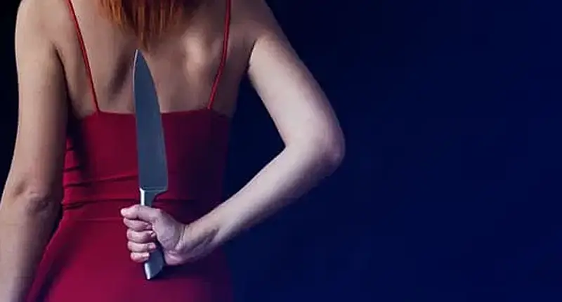 A woman in a red dress holding a knife behind her back with a neutral expression.