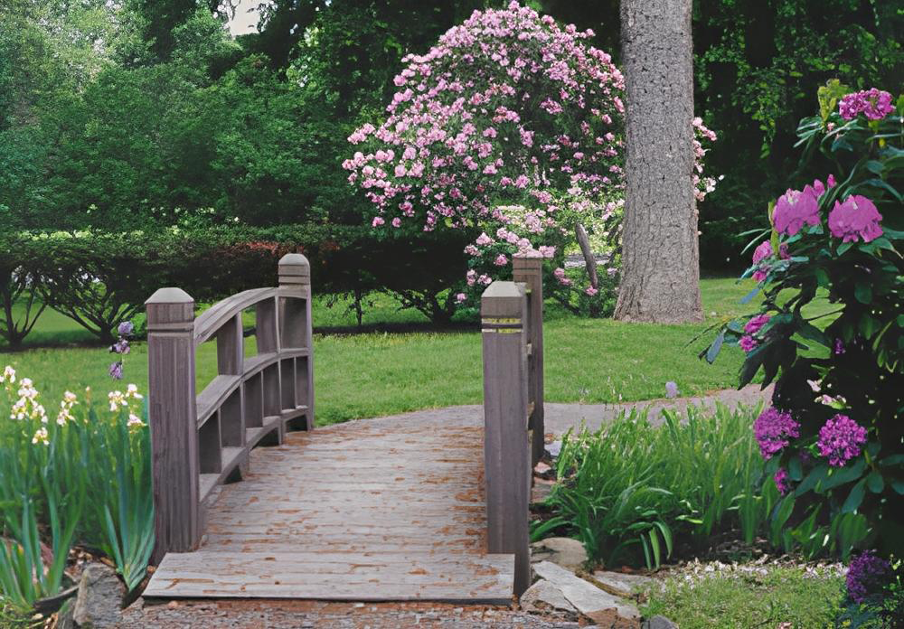 Wooden bridge crossing over a path in a garden with blooming flowers and greenery.