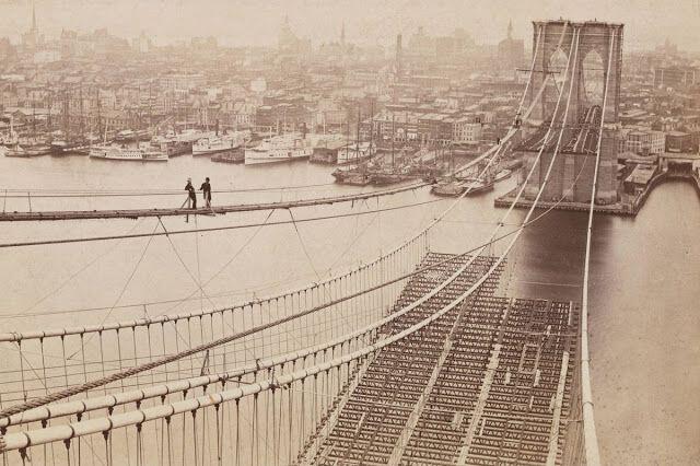 Two workers walking on the cables of the Brooklyn Bridge during its construction in a historical image.