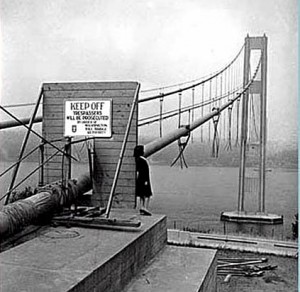 Black and white image of a suspension bridge with a warning sign stating 'Keep Off' in the foreground.