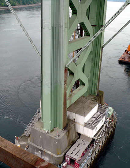 Close-up view of a green bridge support structure with a concrete base and water below, showcasing engineering and construction details.