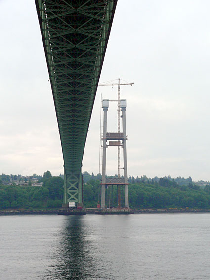 Underneath view of a bridge under construction, showcasing support towers and crane against a cloudy sky.