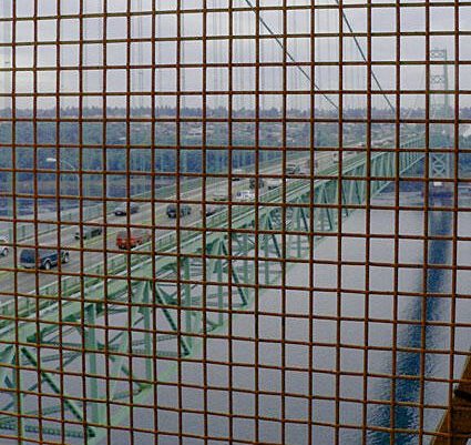 A green suspension bridge viewed through a grid, showcasing vehicles crossing over water, highlighting structural design and engineering.