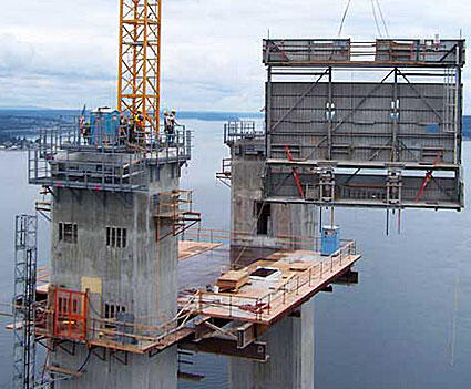 Construction site featuring concrete towers and a crane lifting a large panel over water, showcasing modern building techniques.