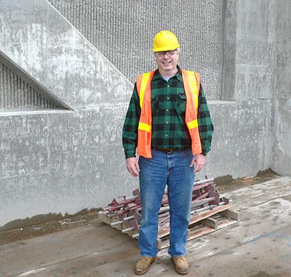 Construction worker wearing a yellow hard hat and orange safety vest standing on a worksite with a concrete wall in the background.