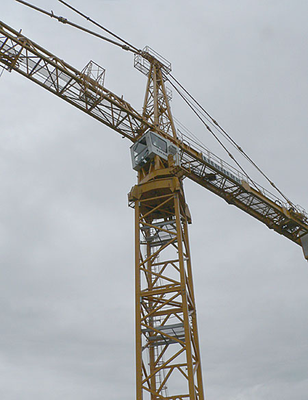 A tall yellow construction crane with a cab and lifting arm against a cloudy sky, used for heavy lifting in construction projects.