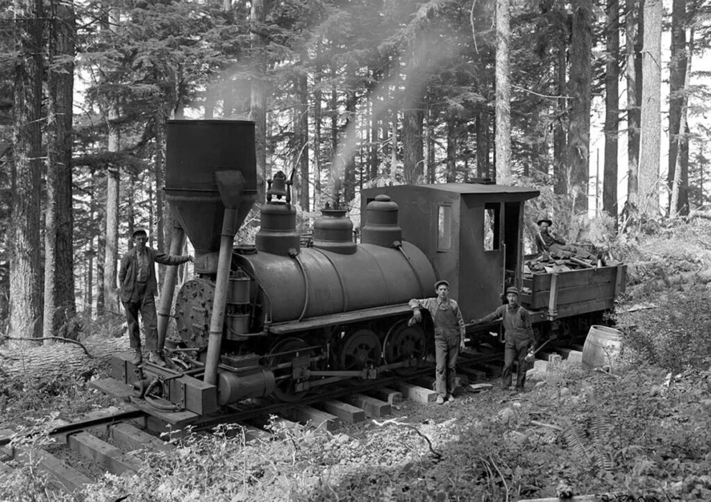 Black and white image of a vintage steam locomotive with workers in a forest setting, showcasing early 20th-century railroad technology.