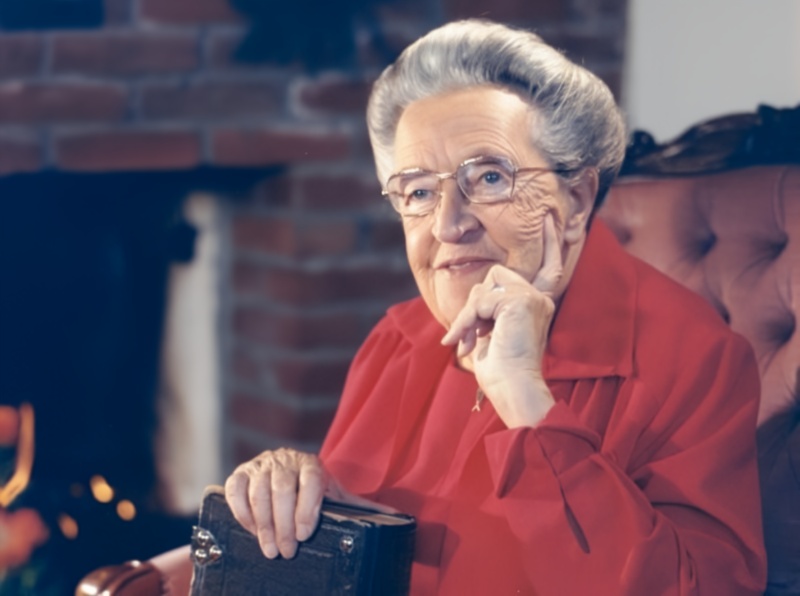 Portrait of an elderly woman with glasses and a red blouse holding a book, sitting in a chair.