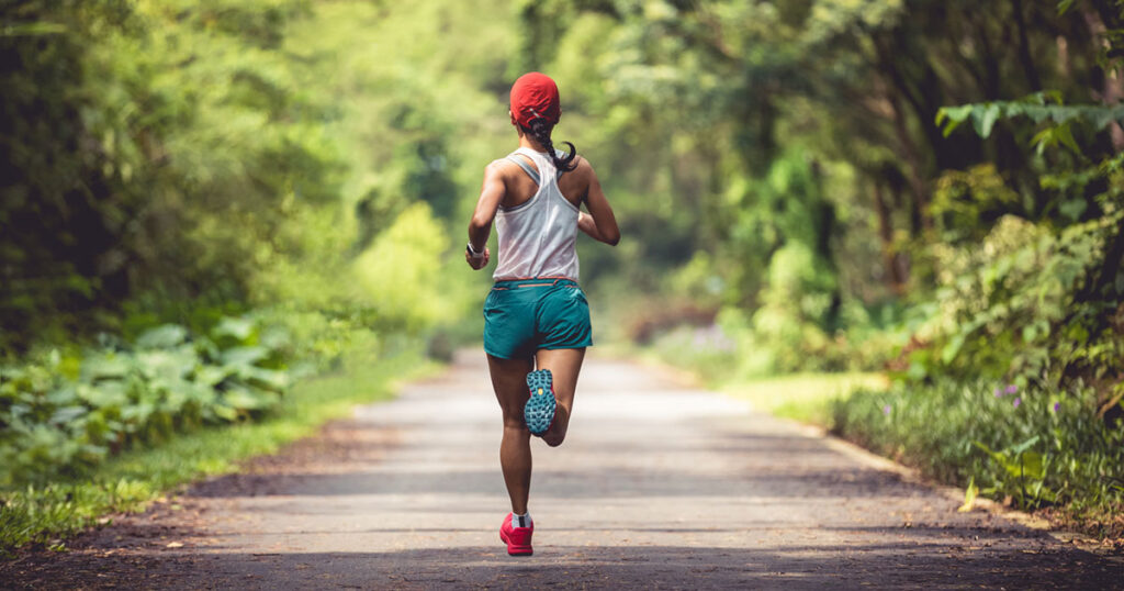 A woman running on a scenic pathway surrounded by greenery, wearing a tank top, shorts, and a red cap, showcasing an active lifestyle.