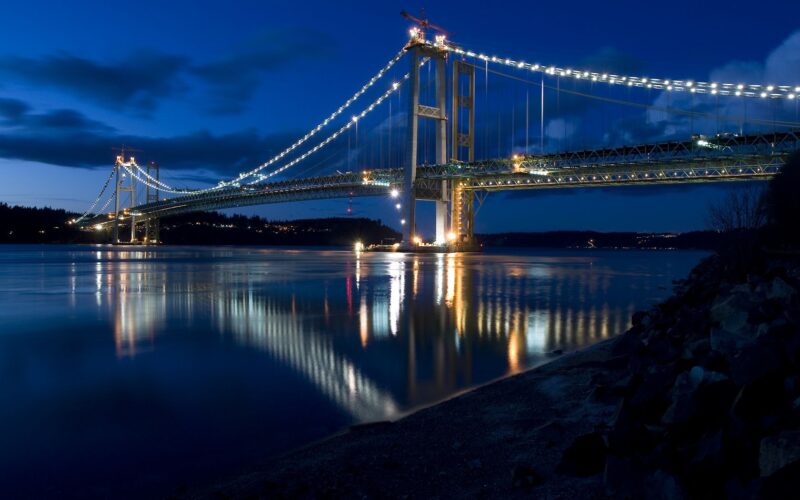 Night view of a newly constructed Tacoma Narrows bridge with lights reflecting on the water, showcasing modern engineering and design.