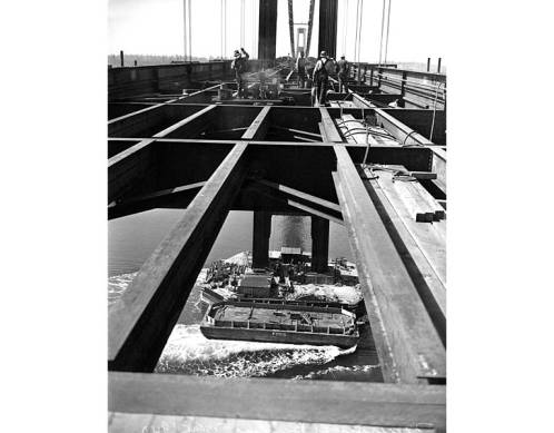 Black and white image of 1st Tacoma Narrows bridge construction with workers on the deck and a barge passing underneath